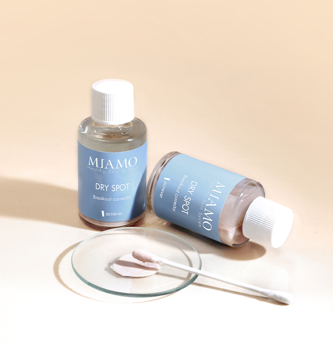 Due flaconcini di DRY SPOT vicini con davanti un vetrino su cui è presente una porzione di prodotto e un cotton fioc. Lo sfondo è beje.